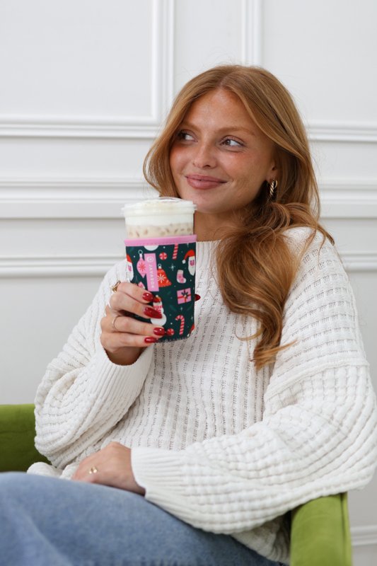 A model in a cream sweater holding an iced drink in a holiday sleeve